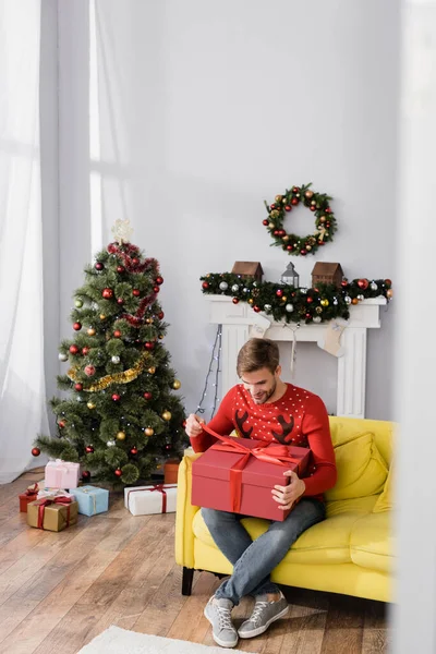 Happy man in red sweater holding wrapped present while sitting on sofa in decorated living room — Stock Photo