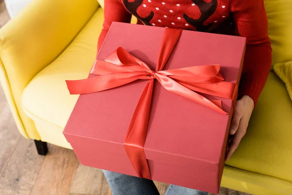 Abgeschnittene Ansicht eines Mannes in rotem Pullover mit eingewickeltem Geschenk, während er auf dem Sofa sitzt mit — Stockfoto