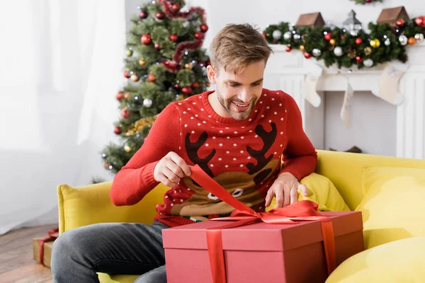Smiling man in red sweater holding ribbon on wrapped present while sitting on sofa with blurred christmas tree on background — Stock Photo