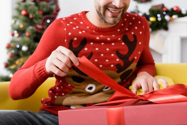 Обрезанный вид веселого человека в красном свитере, держащего ленточку на подарок, сидя на диване — стоковое фото