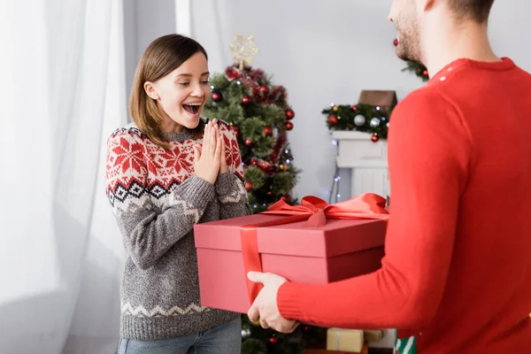 Mann im roten Pullover hält umwickeltes Geschenk neben aufgeregter Frau und Weihnachtsbaum auf verschwommenem Hintergrund — Stockfoto