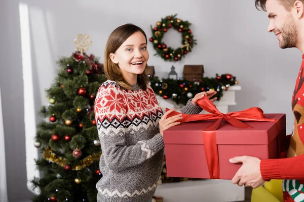 Cheerful man in red sweater holding wrapped present near happy wife and christmas tree on blurred background — Stock Photo