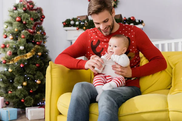 Felice padre che alimenta il figlio neonato mentre tiene il biberon con latte materno vicino all'albero di Natale su sfondo sfocato — Foto stock