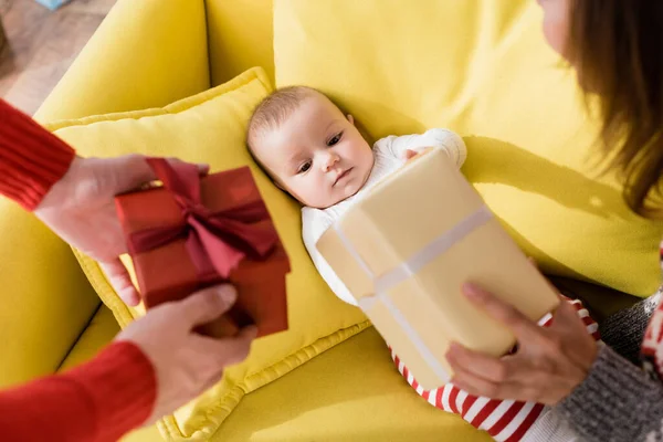 Младенец сын лежит на диване и смотрит на родителей, держащих завернутые рождественские подарки на размытом переднем плане — стоковое фото