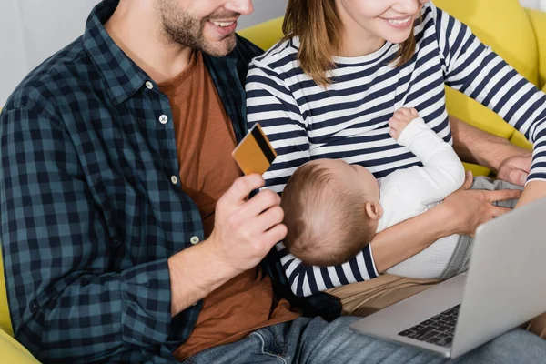 Homem alegre segurando cartão de crédito perto da esposa com filho bebê e laptop — Fotografia de Stock