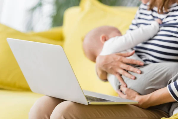 Laptop on knees of mother holding in arms baby boy on blurred background — Stock Photo
