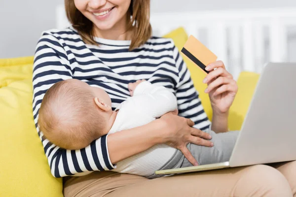 Vue recadrée d'une femme souriante avec un fils en bas âge tenant une carte de crédit près d'un ordinateur portable assis sur un canapé — Photo de stock