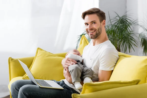 Feliz padre sosteniendo en brazos sueño bebé hijo mientras está sentado en el sofá con el ordenador portátil - foto de stock