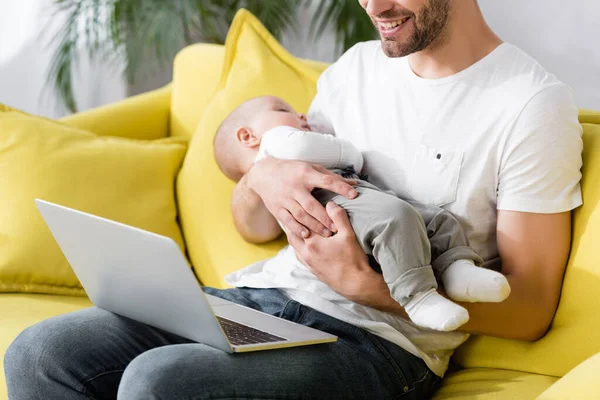 Alegre padre sosteniendo en brazos somnoliento bebé hijo mientras está sentado en sofá con portátil - foto de stock