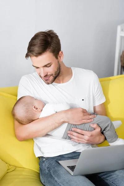 Père attentionné tenant dans les bras bébé fils endormi tout en étant assis sur le canapé avec ordinateur portable — Photo de stock