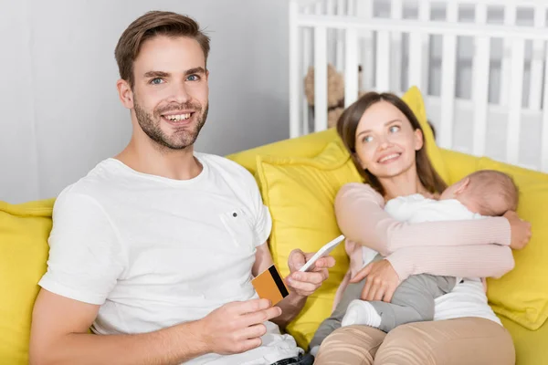 Cheerful man holding credit card and smartphone near wife with infant son on blurred foreground — Stock Photo