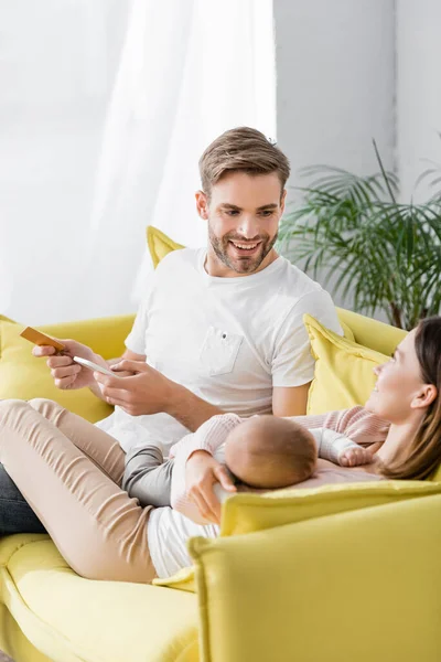 Hombre feliz sosteniendo tarjeta de crédito y teléfono inteligente cerca de la esposa con hijo pequeño en primer plano borrosa - foto de stock