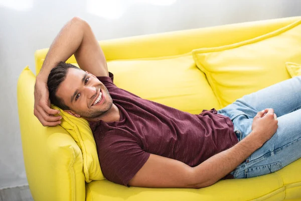 Fröhlicher Mann blickt in Kamera, während er zu Hause auf gelbem Sofa liegt — Stockfoto