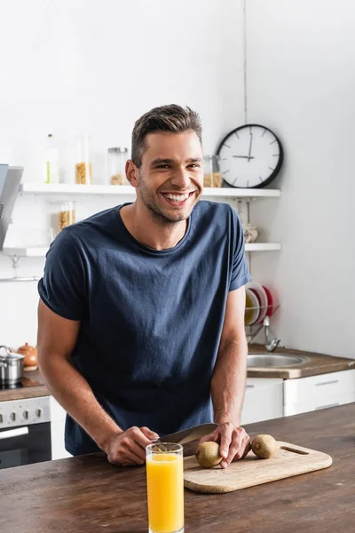 Homem sorridente segurando faca perto de kiwi na placa de corte e vidro de suco de laranja na mesa — Fotografia de Stock