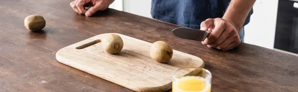 Vista cortada do homem segurando faca perto de kiwi na placa de corte perto de vidro de suco de laranja em primeiro plano borrado, banner — Fotografia de Stock