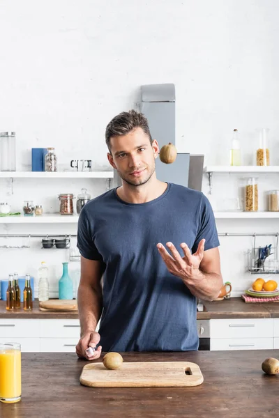 Young man throwing kiwi and holding knife near glass of orange juice and cutting board in kitchen - foto de stock