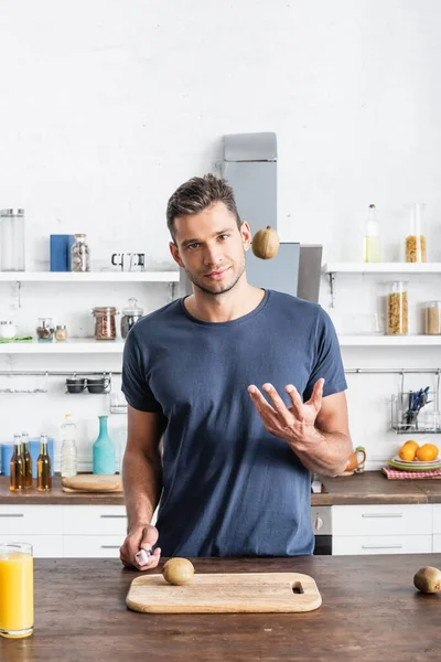 Jovem jogando kiwi e segurando faca perto de vidro de suco de laranja e tábua de corte na cozinha — Fotografia de Stock