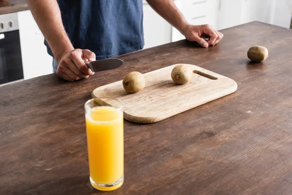 Cropped view of man holding knife near kiwi on cutting board and glass of orange juice on blurred foreground - foto de stock