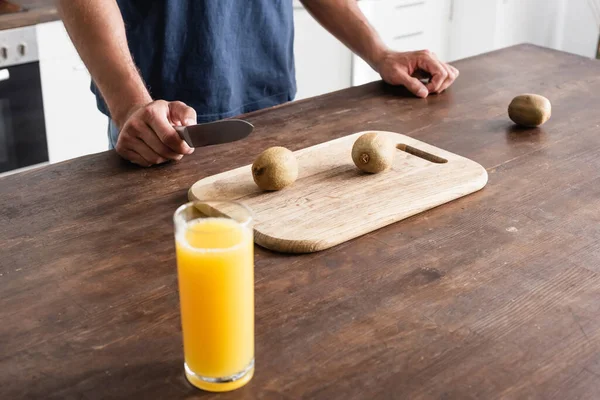 Abgeschnittene Ansicht eines Mannes mit Messer in der Nähe von Kiwi auf Schneidebrett und Glas Orangensaft im unscharfen Vordergrund — Stockfoto