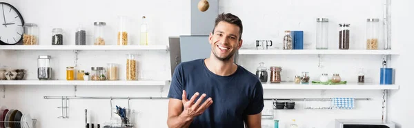 Smiling man throwing kiwi and looking at camera in kitchen, banner - foto de stock