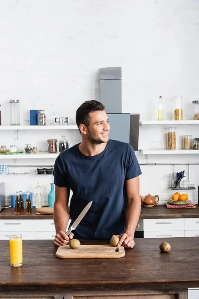 Smiling man with knife holding kiwi near cutting board and orange juice on table — Stock Photo