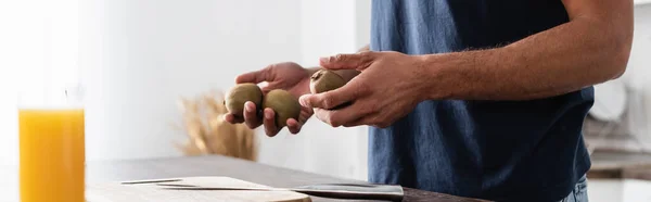 Cropped view of man holding kiwi near knife, cutting board and orange juice on blurred foreground, banner - foto de stock