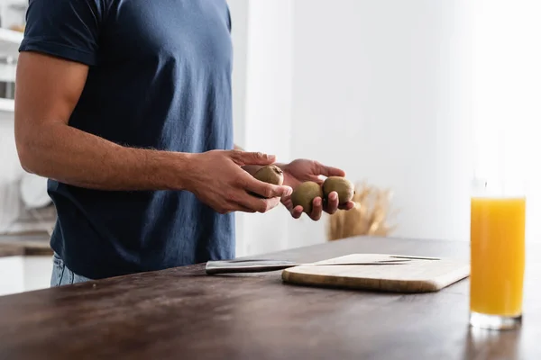 Cropped view of man holding fresh kiwi near cutting board, knife and glass of orange juice on blurred foreground — Stock Photo