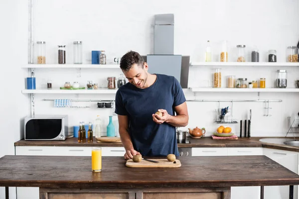 Улыбающийся мужчина смотрит на киви на разделочной доске возле апельсинового сока в стекле на столе на кухне — стоковое фото
