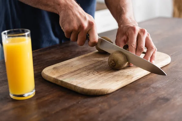 Vista cortada do homem que corta o kiwi perto do vidro do suco de laranja no primeiro plano borrado — Fotografia de Stock