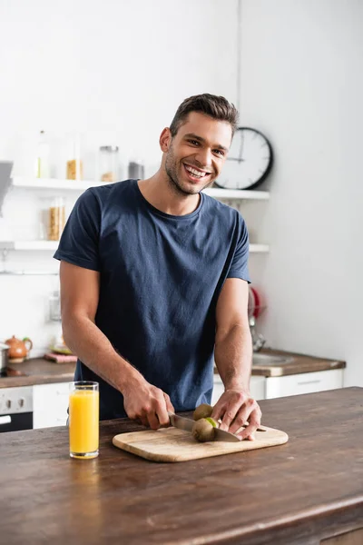 Lächelnder Mann blickt in die Kamera, während er Kiwi auf Schneidebrett in der Nähe von Orangensaft in der Küche schneidet — Stockfoto