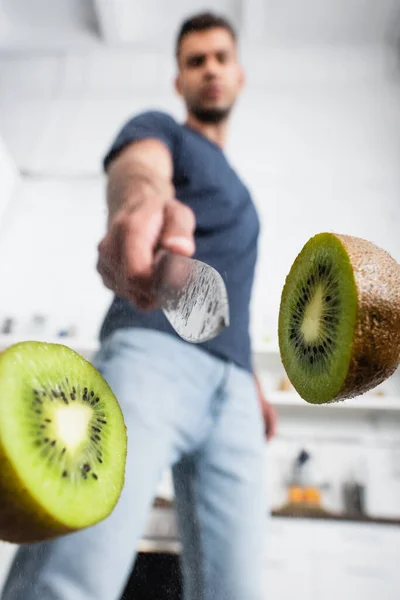 Metades de kiwi fresco perto da faca na mão do homem no fundo borrado — Fotografia de Stock