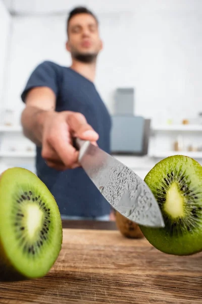 Visão de perto de metades de kiwi suculento perto de faca em gotas de água na mão do homem no fundo borrado — Fotografia de Stock