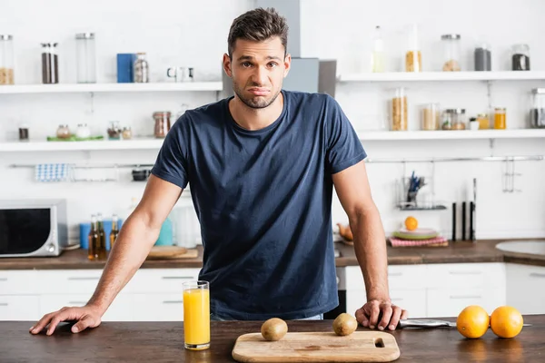 Trauriger Mann blickt in Kamera neben Orangensaft, Früchten und Schneidebrett auf Tisch — Stockfoto