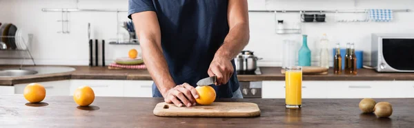 Vista ritagliata dell'uomo che taglia arancia fresca vicino al kiwi e vetro di succo d'arancia, banner — Foto stock