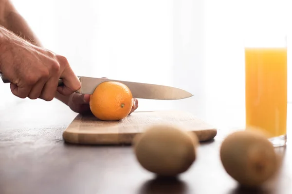 Vista ritagliata dell'uomo che taglia l'arancia vicino al kiwi e al bicchiere di succo d'arancia in primo piano sfocato — Foto stock