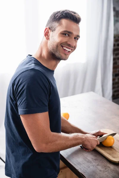 Vista lateral do homem sorrindo para a câmera ao cortar laranja no fundo embaçado na cozinha — Fotografia de Stock