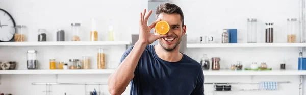 Uomo sorridente che tiene mezza arancia fresca vicino al viso in cucina, banner — Foto stock