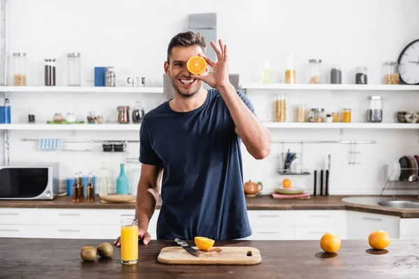 Cheerful man holding half of orange near face beside kiwi, cutting board and orange juice in kitchen — Stock Photo