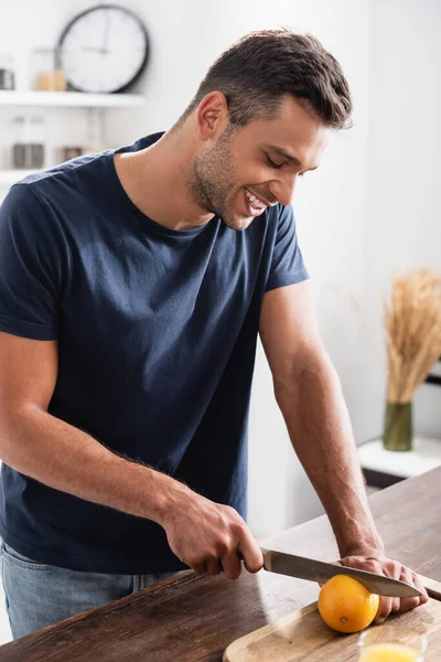 Homme joyeux coupant orange près du verre de jus sur le premier plan flou — Photo de stock