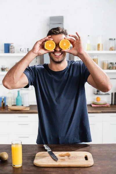 Young man smiling while covering face with halves of orange near cutting board and orange juice in kitchen — Stock Photo