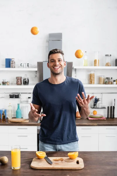 Cheerful man throwing oranges near cutting board and glass of orange juice — Stock Photo