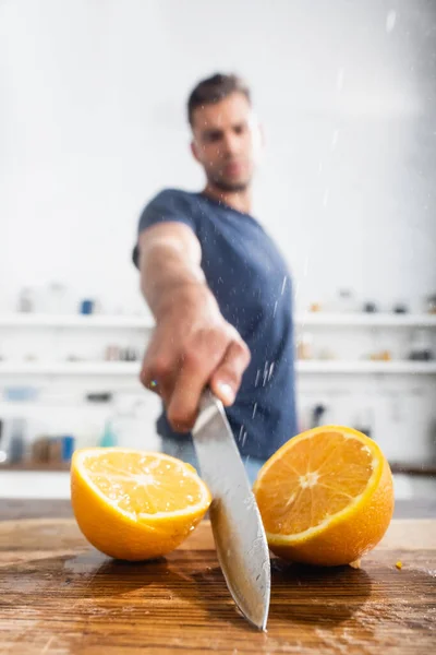 Close up view of halves of orange and wet knife in hand of man on blurred background — Stock Photo