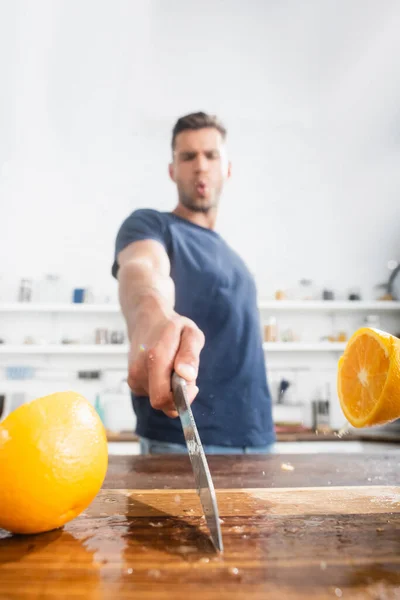 Close up view of halves of orange on wet cutting board and knife in hand of young man on blurred background - foto de stock