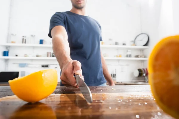 Cropped view of knife on wet cutting board near halves of orange and man on blurred background — Stock Photo