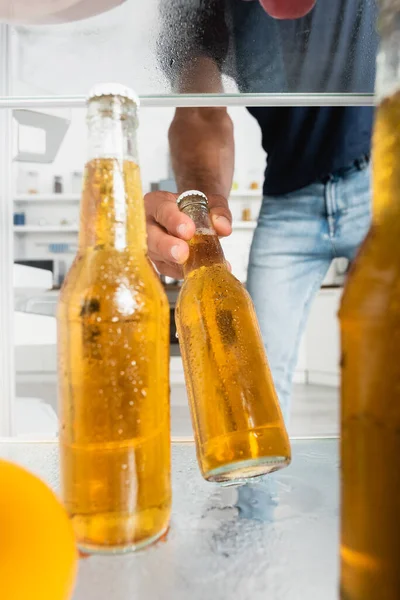 Влажная бутылка пива в руке человека рядом с открытым холодильником на размытом фоне — стоковое фото