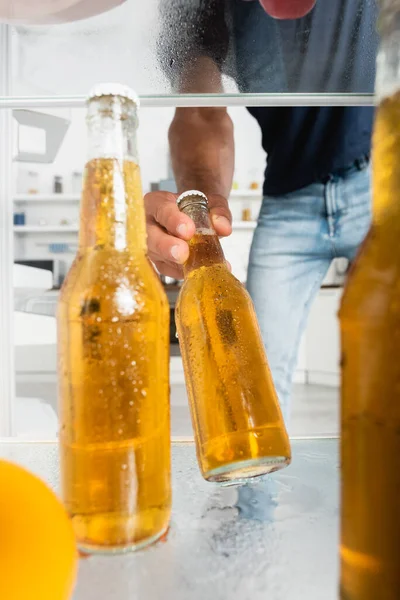 Bottiglia bagnata di birra in mano dell'uomo vicino al frigorifero aperto su sfondo sfocato — Foto stock