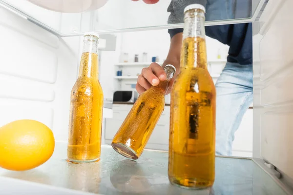 Обрезанный вид человека, берущего влажную бутылку пива возле апельсина на размытом переднем плане в холодильнике — стоковое фото