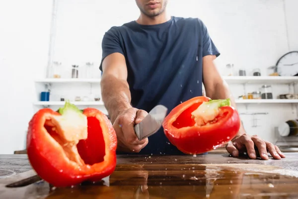 Cropped view of man holding knife near halves of fresh bell pepper on cutting board on blurred foreground — Stock Photo