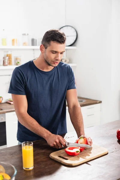 Young man cutting paprika near glass of orange juice on blurred foreground in kitchen - foto de stock