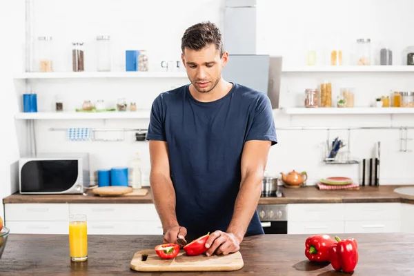 Young man cutting bell pepper near glass of orange juice in kitchen - foto de stock